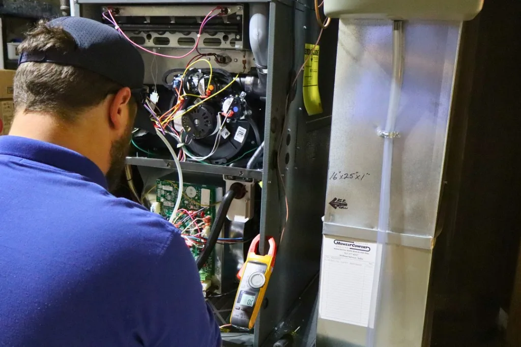 A Midwest Comfort technician in a blue polo carefully inspecting the internals of an open residential furnace during a Comfort Club tune-up visit